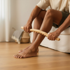Homme assis au bord du canapé qui fait rouler le bâton de massage en hêtre sur son mollet, vue basse au ras du sol