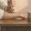 Marteau d'acupression en métal appuyé sur l'avant-bras posé sur une table en noyer, main d'une femme sur le manche, lumière chaude de l'après-midi
